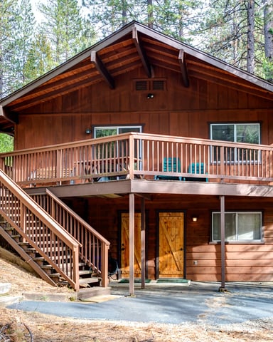 Two-story wood cabin with wraparound deck surrounded by tall pine trees in forest setting