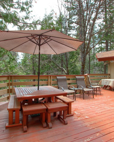 Wooden deck with dining table, lounge chairs, umbrella, and cabin surrounded by tall pine trees