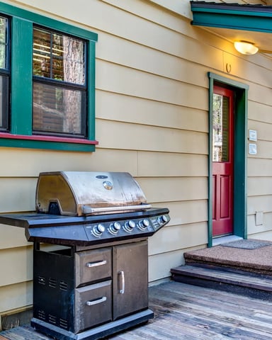 Cabin exterior with gas grill on deck, teal-trimmed windows and red door, forest visible in background