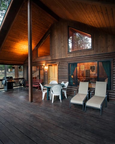 Wide covered deck on a log A-frame cabin with Adirondack chairs, grill, and forest views