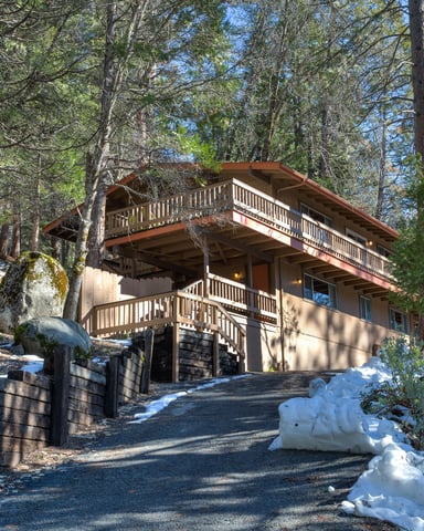 Two-story cabin with wooden railings nestled among tall pine trees, with patches of snow on the ground and driveway.
