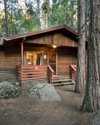 Classic log cabin with stone chimney glowing at dusk, tucked among boulders and tall redwoods