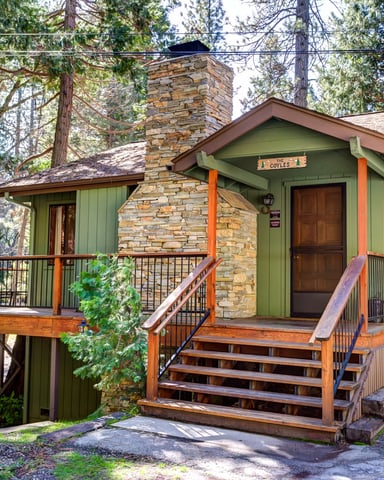 Green cabin with stone chimney, wood deck, and stairs surrounded by tall pine trees