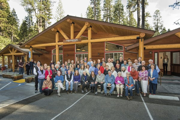 Large group of people gathered outside a log cabin lodge with tall timber columns, surrounded by pine forest