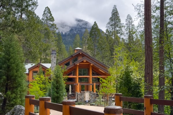 Log cabin with stone chimney surrounded by tall pine trees and mountain peaks with low clouds