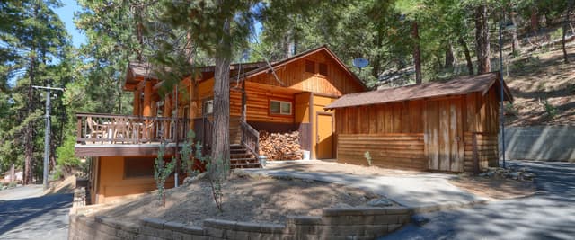 Wood cabin with dark roof nestled among tall pine trees, featuring a wraparound deck and native vegetation in foreground.