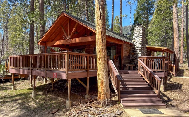 Log cabin with stone chimney and wraparound deck among tall pines in Wawona