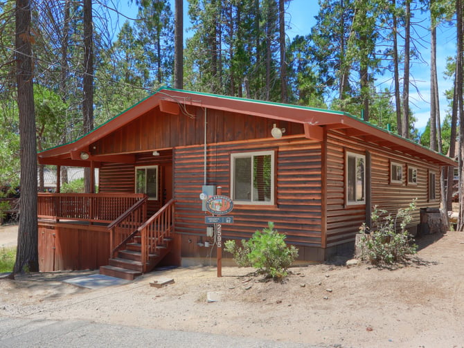 Red-brown log cabin with green roof and front porch surrounded by tall pine trees