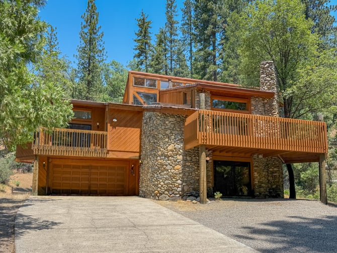 Modern wood and stone cabin with multiple decks surrounded by tall pine trees and clear blue sky