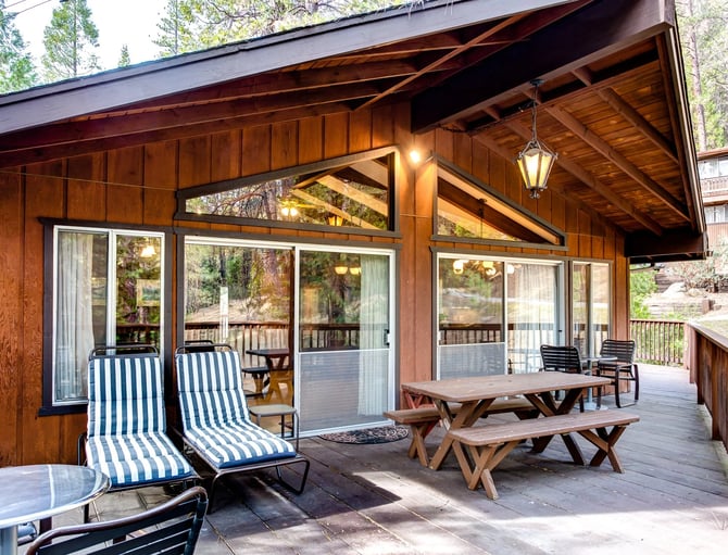 Wooden cabin deck with striped lounge chairs, picnic tables, and glass doors overlooking forest